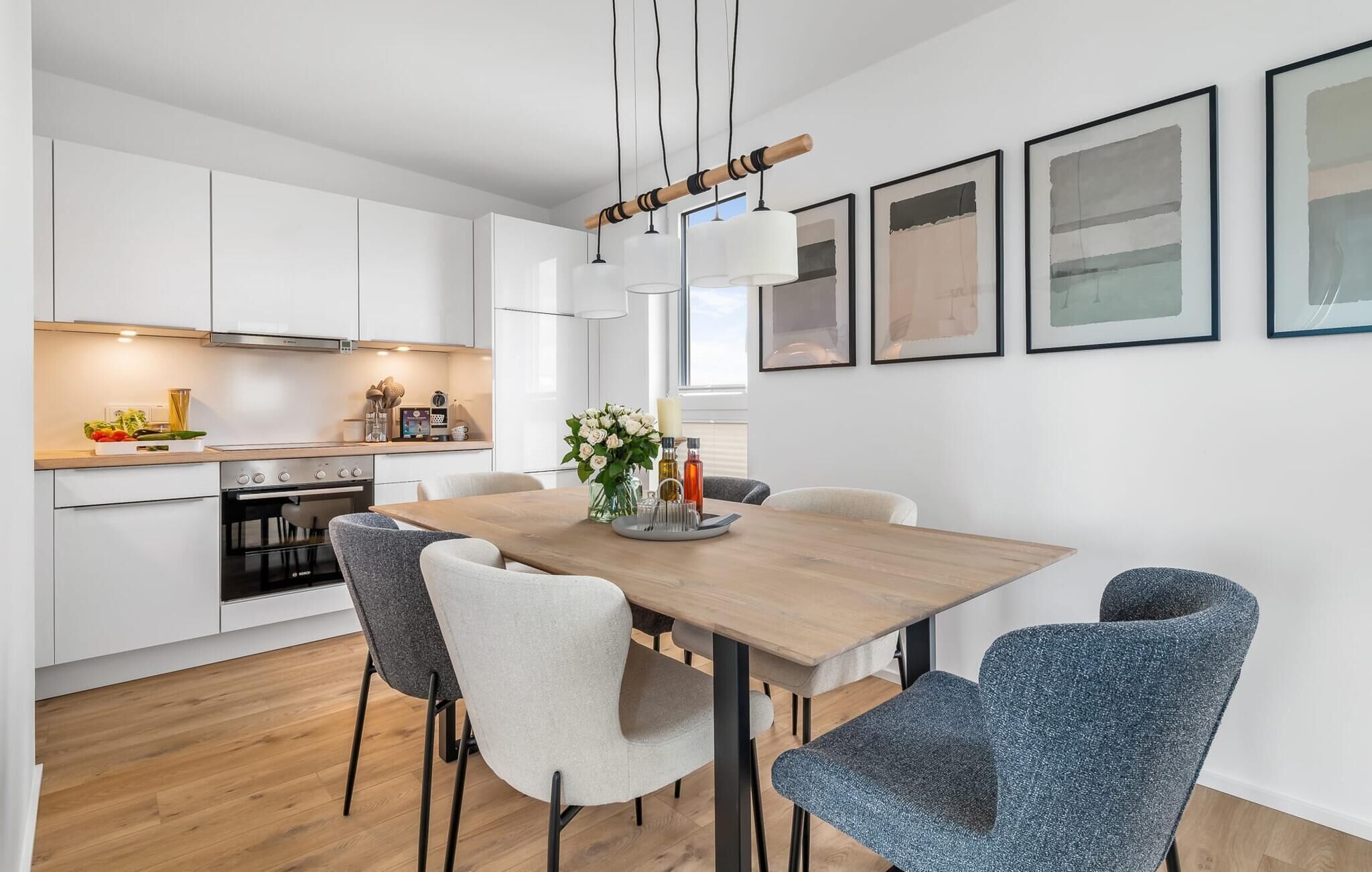 Une cuisine moderne avec des meubles blancs, une table à manger en bois et des chaises grises. Des œuvres d’art encadrées ornent le mur, et un luminaire suspendu se trouve au-dessus de la table.