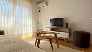 Une chambre d’hôtel moderne, équipée d’un lit, d’un téléviseur posé sur un meuble en bois, d’une petite table avec une chaise et d’une grande fenêtre ornée de rideaux légers.