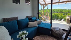 Een moderne woonkamer met een blauwe modulaire bank, een klein rond tapijt en uitzicht op een balkon met buitenmeubilair.