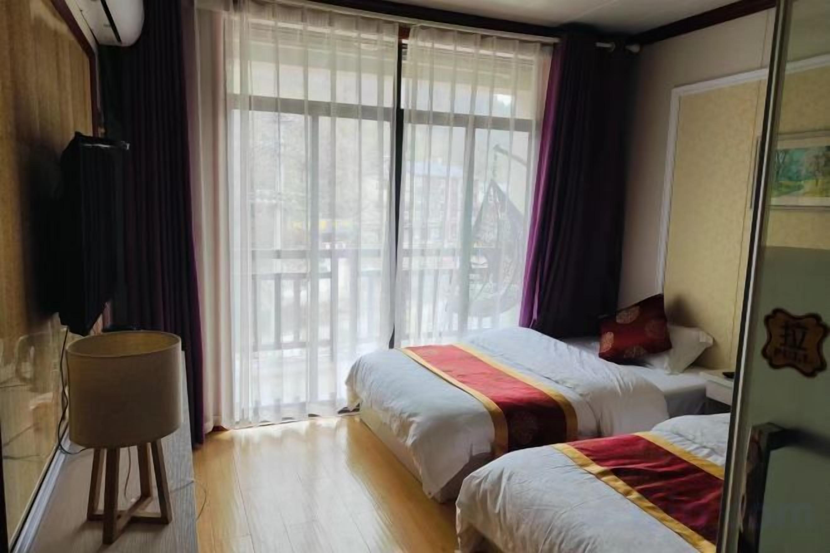 A hotel room with two beds, a large window with curtains, a bedside table, and a wall-mounted TV.