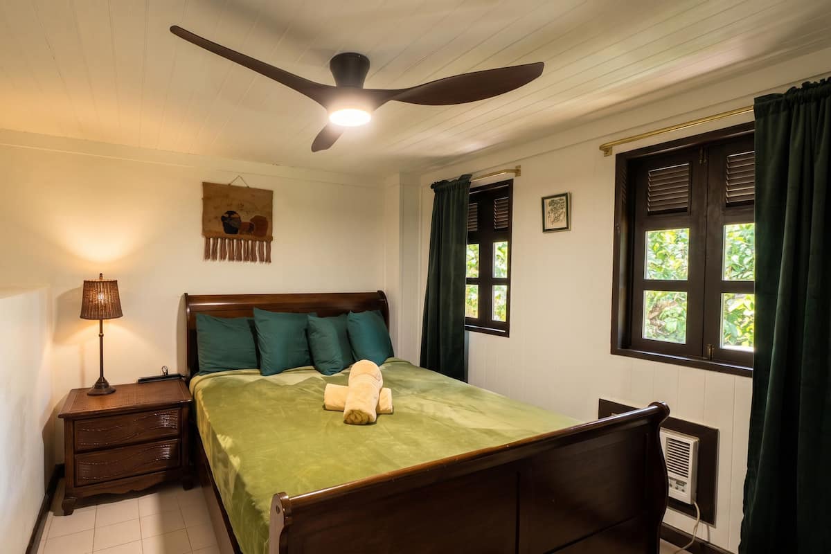 A bedroom with a bed, a nightstand with a lamp, a ceiling fan, a window with curtains, and a wall-mounted air conditioning unit.