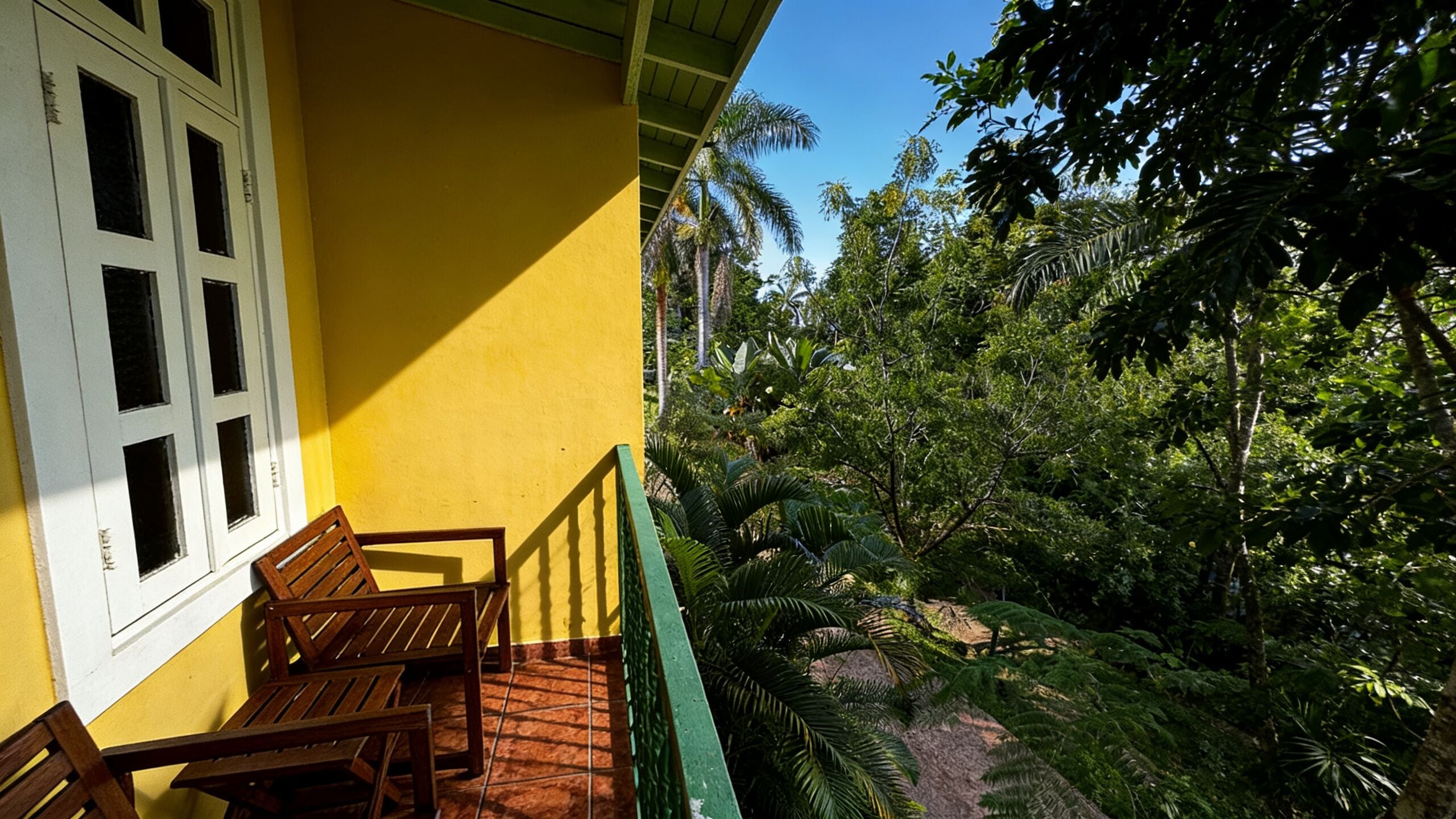 Un bâtiment jaune doté d’un balcon, de chaises en bois et offrant une vue sur un feuillage verdoyant et luxuriant.
