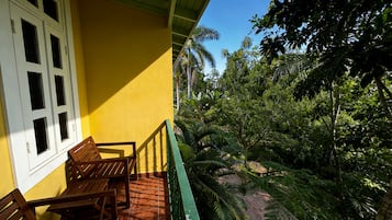 Un bâtiment jaune doté d’un balcon, de chaises en bois et offrant une vue sur un feuillage verdoyant et luxuriant.