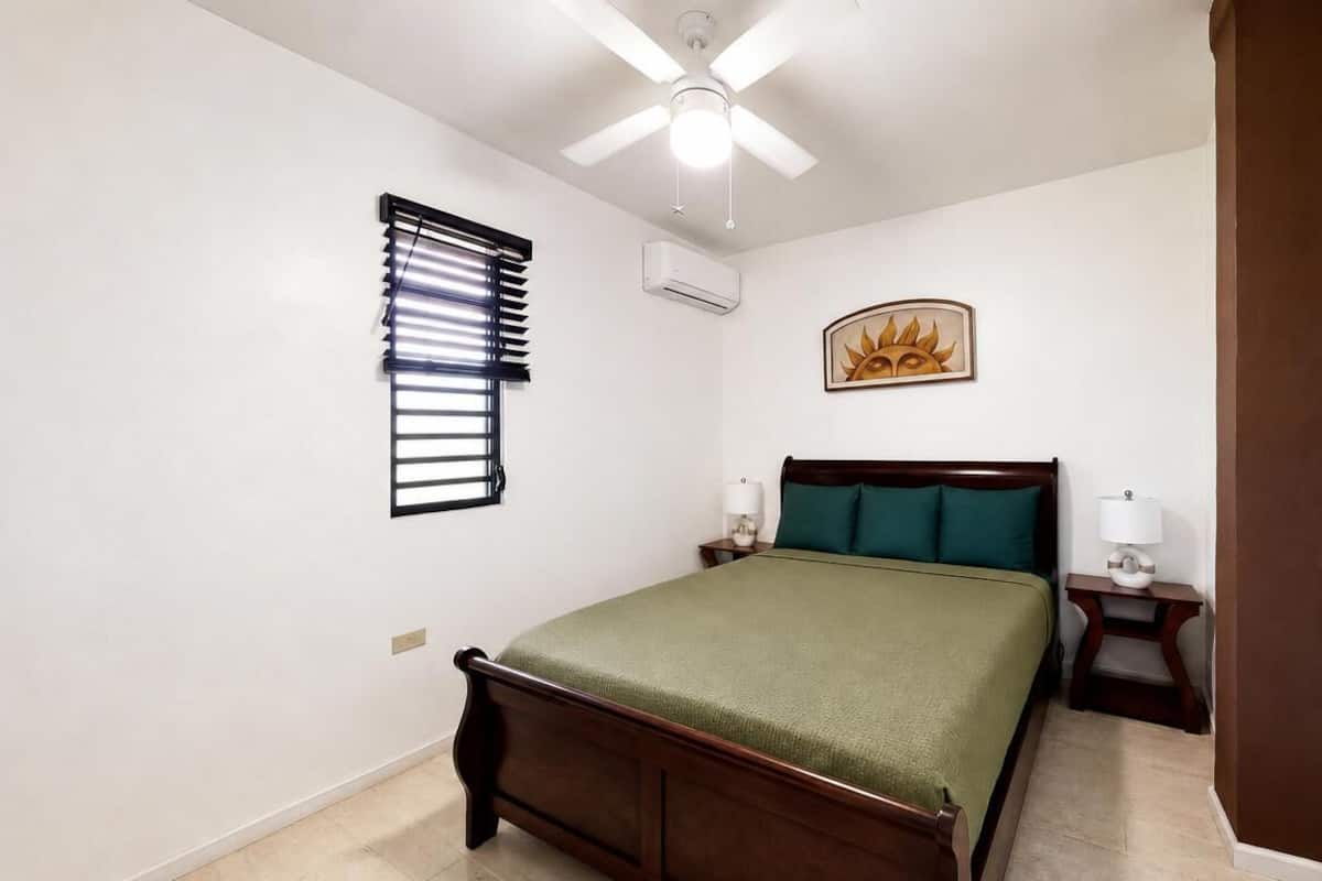 A bedroom with a bed, a ceiling fan, an air conditioning unit, a window with blinds, a bedside table with a lamp, and a wall-mounted picture.