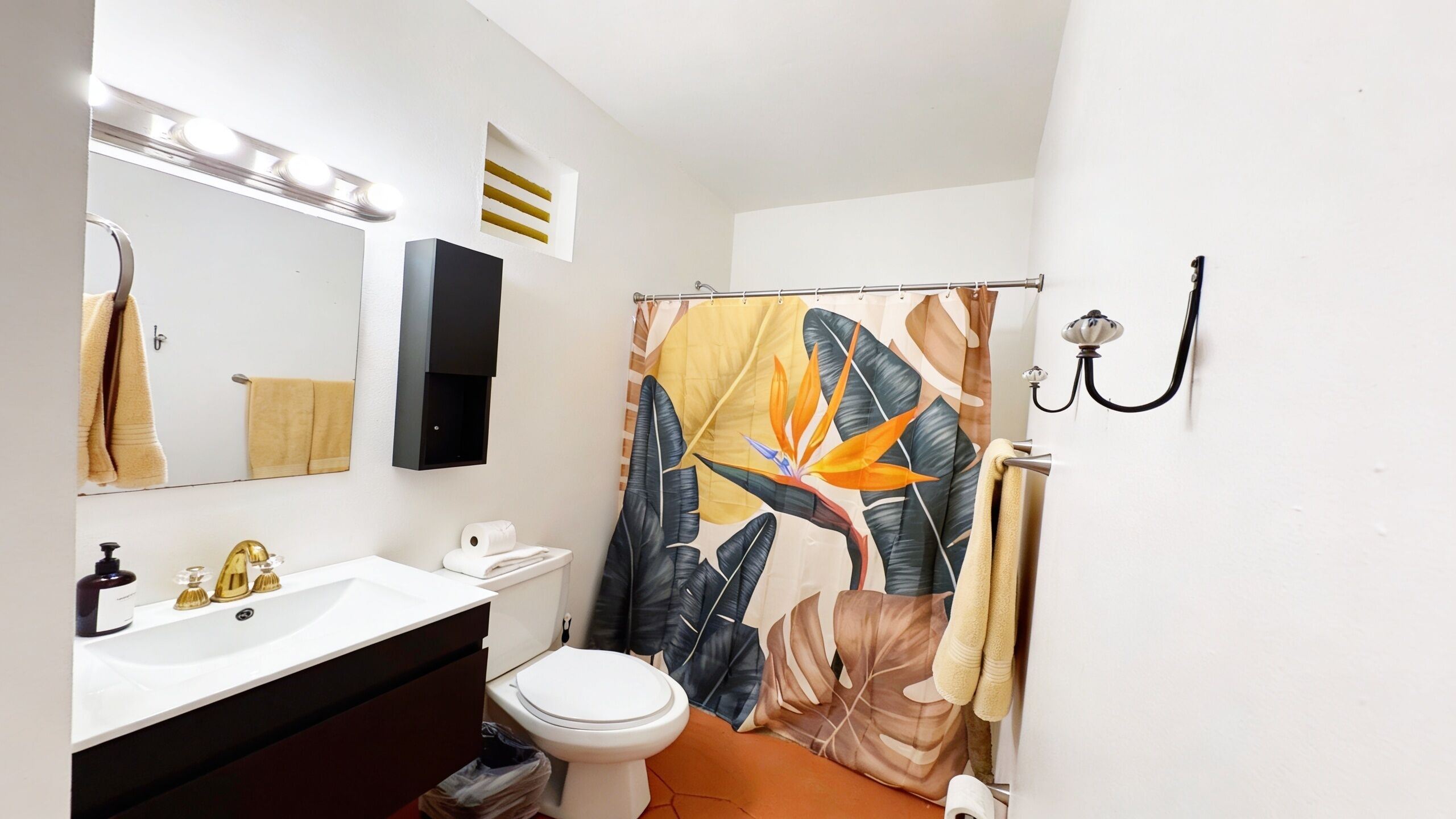 Un baño con un lavamanos blanco, un inodoro, una cortina de ducha con estampado de hojas tropicales, un perchero y una lámpara fijada a la pared.