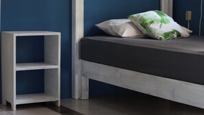 A bed with a grey mattress and white pillows, a white bedside table, and a blue wall. - Experiencia Surf Camp (Brisas de Zicatela)