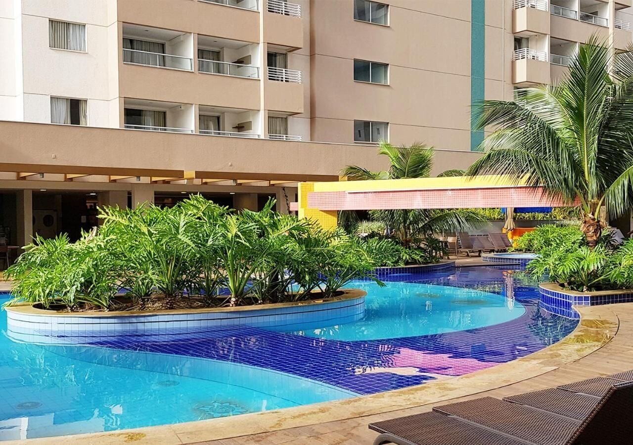 A swimming pool with tiled edges, surrounded by greenery and a building with balconies in the background.