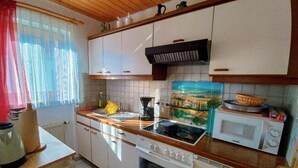 A kitchen with white cabinets, a stove, a microwave, and a window with sheer curtains. - Schöne, große, barrierefreie Ferienwohnung, ruhig, idyllisch, familienfreundlich (Todtmoos)