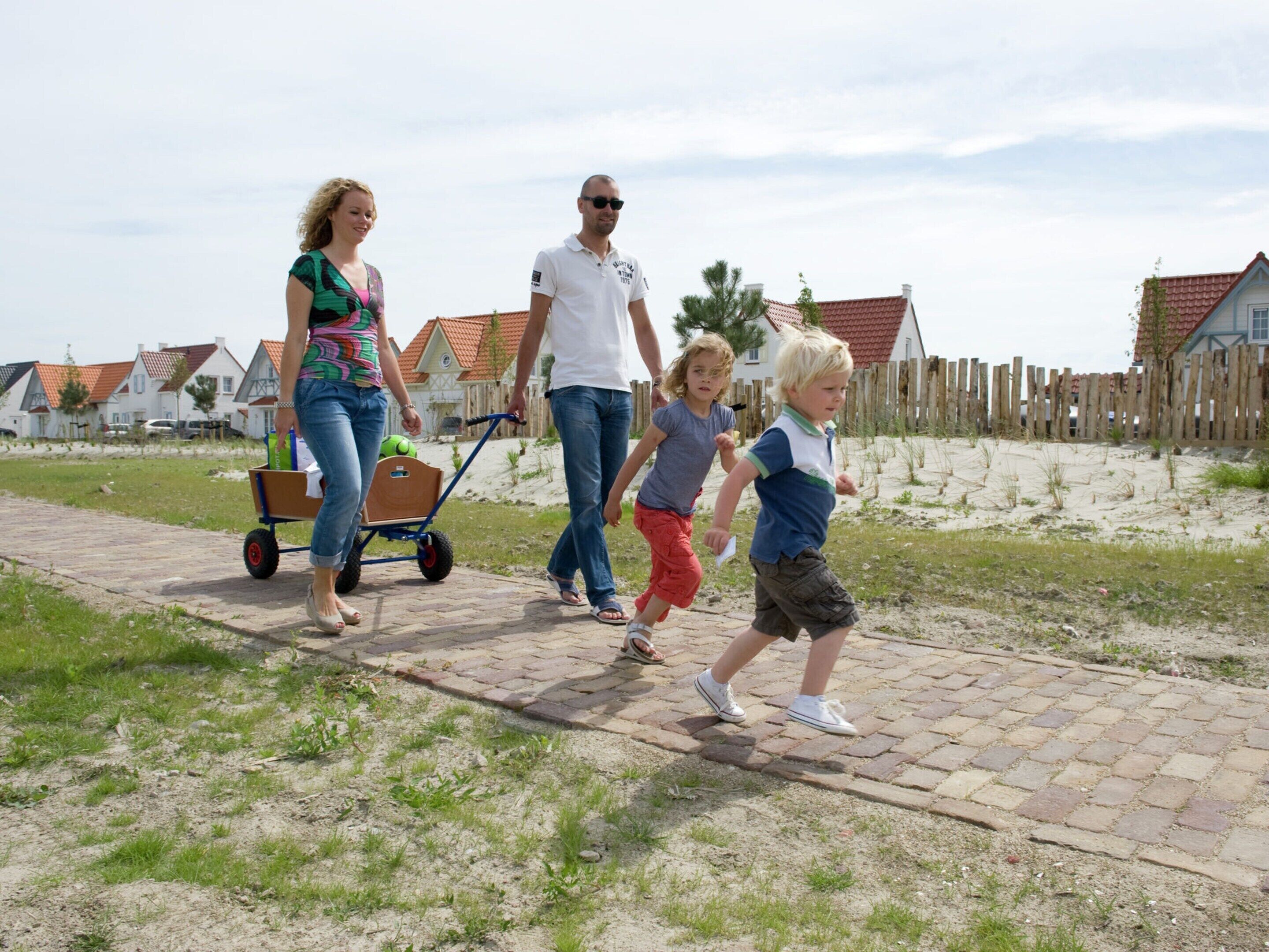 A family walking on a paved path with a wooden cart, surrounded by houses and a fence.