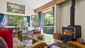 A cozy living room with a fireplace, wooden furniture, and large windows offering a view of greenery. - Azimuth – Family Retreat with Sauna, Garden & Scenic Views (Wentworth Falls)