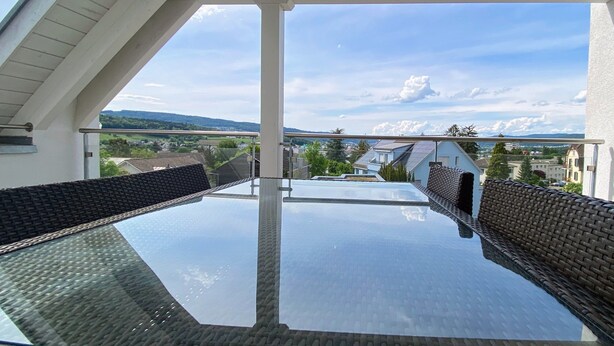 A glass-top table with wicker furniture, a clear view of a mountainous landscape, and a white ceiling structure.