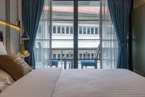 A hotel room with a large bed, a nightstand, a lamp, and a view of a building through a large window with blue curtains. - Momo Hotel (Chiang Mai)