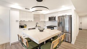 Een moderne keuken met witte kasten, een marmeren eiland, groene stoelen, roestvrijstalen apparatuur en een hanglamp.