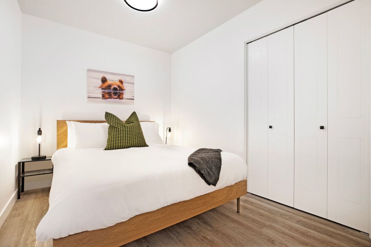 A bedroom with a wooden bed, white bedding, a green pillow, a gray throw, a nightstand with a lamp, a wall-mounted picture, and a white wardrobe.
