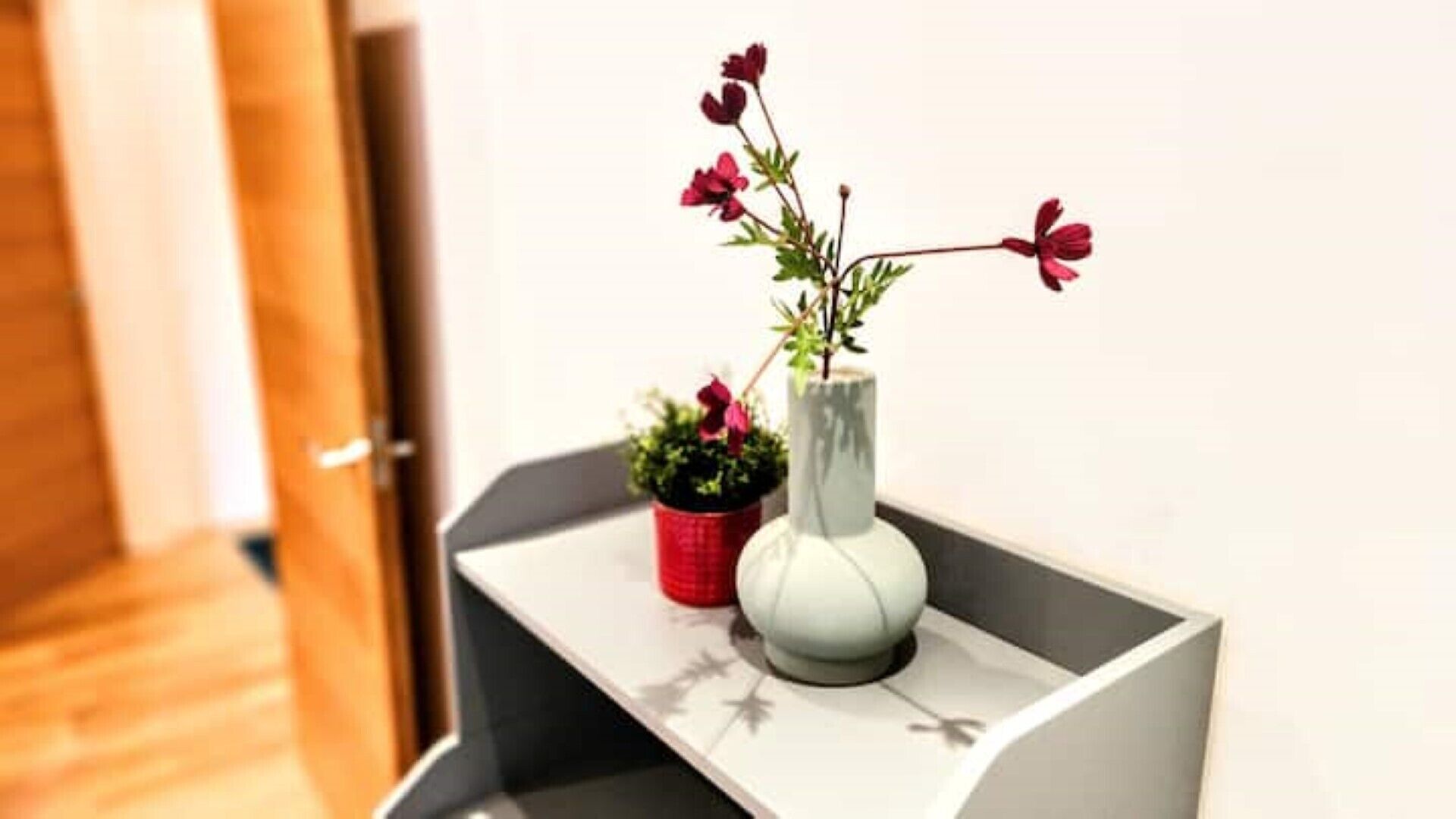 A white vase with red flowers on a white shelf.