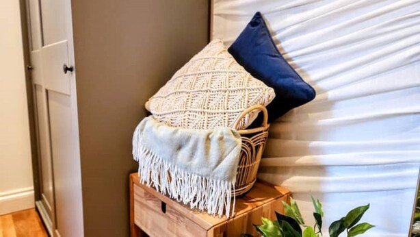 Rincón de una habitación con una cesta tejida, una almohada azul y una manta blanca con flecos sobre un banco de madera.