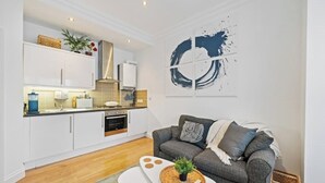 Cocina moderna con armarios blancos, horno integrado y zona de comedor con sofá gris y una mesita.