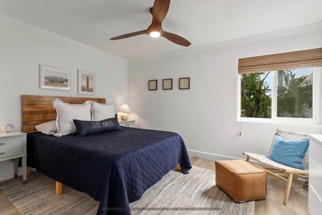 A bedroom with a bed, a nightstand, a chair, a window with a view of greenery, and a ceiling fan.
