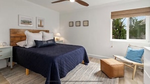 A bedroom with a bed, a nightstand, a chair, a window with a view of greenery, and a ceiling fan.