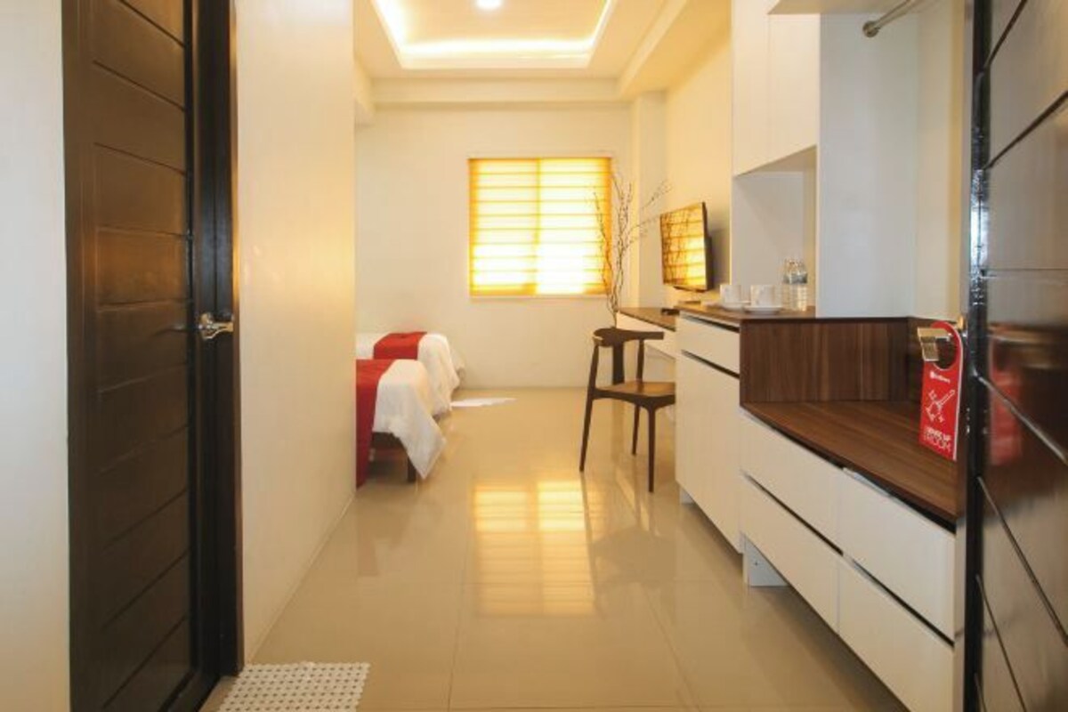 A hotel room with a kitchenette, a bed with a red headboard, a window with blinds, and a small table with a chair.