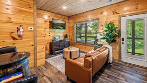 A wooden cabin interior with a leather couch, a round coffee table, a fireplace, a window with a view, and a fire extinguisher on the wall. - Smoky Mountain Getaway • 2 King Suites • Hot Tub • Near Dollywood • Pets (Sevierville)