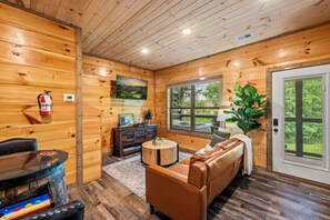 A wooden cabin interior with a leather couch, a round coffee table, a fireplace, a window with a view, and a fire extinguisher on the wall.