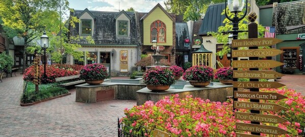 A quaint village square with cobblestone paths, flower beds, and wooden signposts pointing to various establishments.