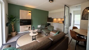 Una sala moderna con un sofá, sillas, una mesita y un televisor. Se observa una zona de cocina con armarios y electrodomésticos a través de una puerta abierta.
