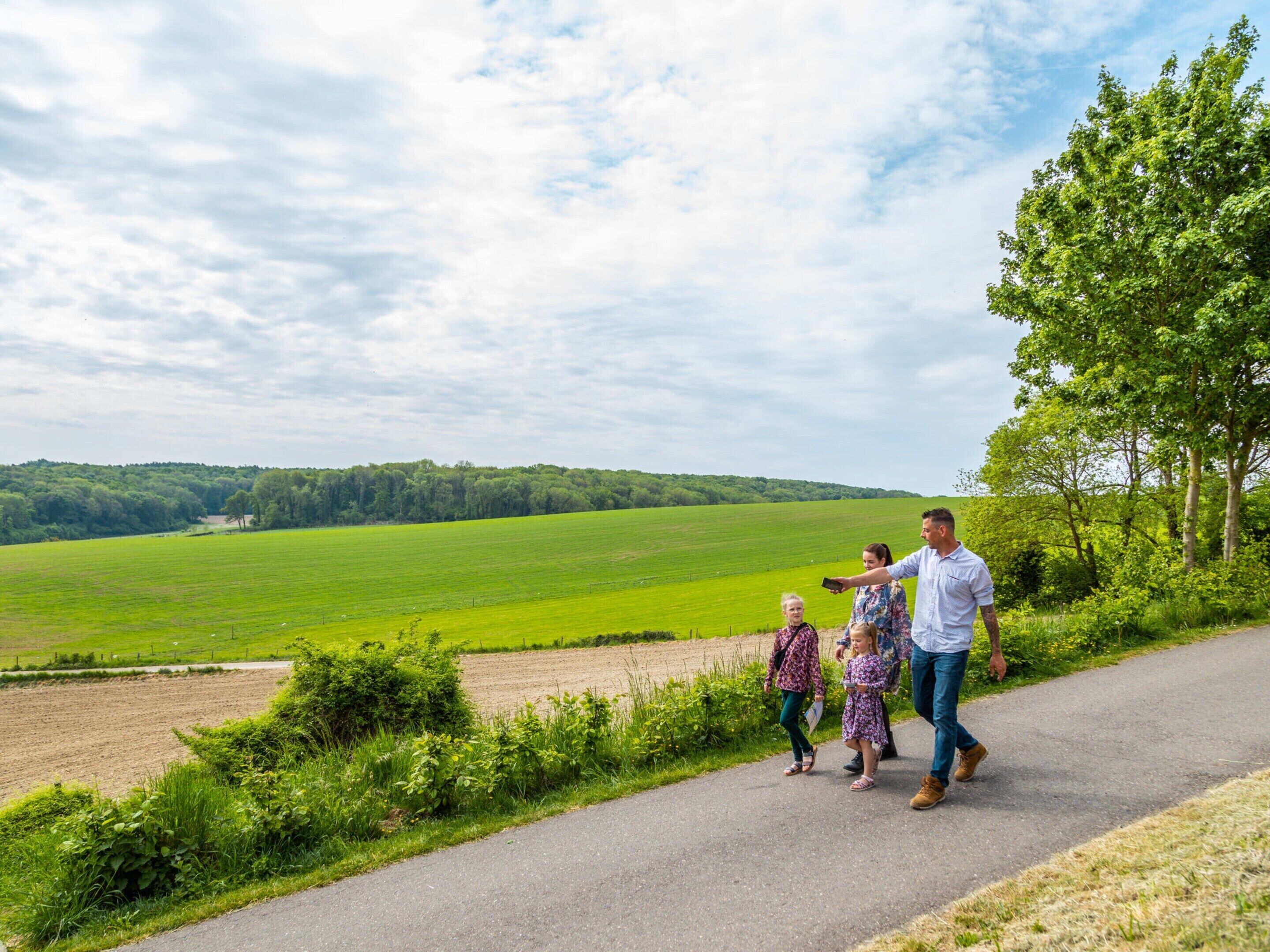 Une famille se promène sur un chemin pavé, le long d’un champ verdoyant, avec un arbre sur la droite.