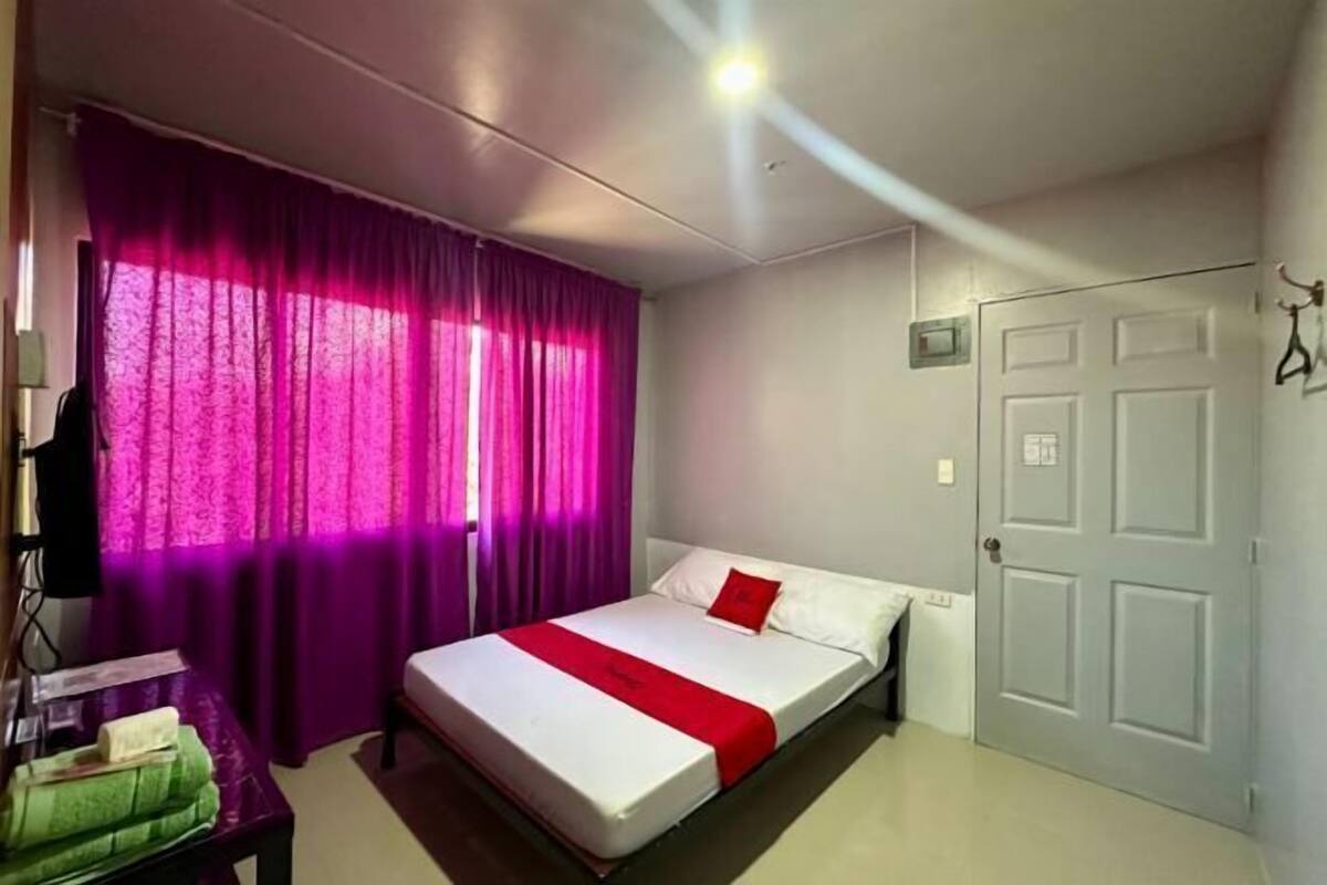 A bedroom with a bed, a window with purple curtains, a door, and a small table with a green towel.