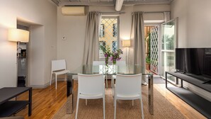 Un salon moderne avec une table à manger en verre, des chaises blanches, un canapé noir, un téléviseur et une grande fenêtre avec des rideaux.