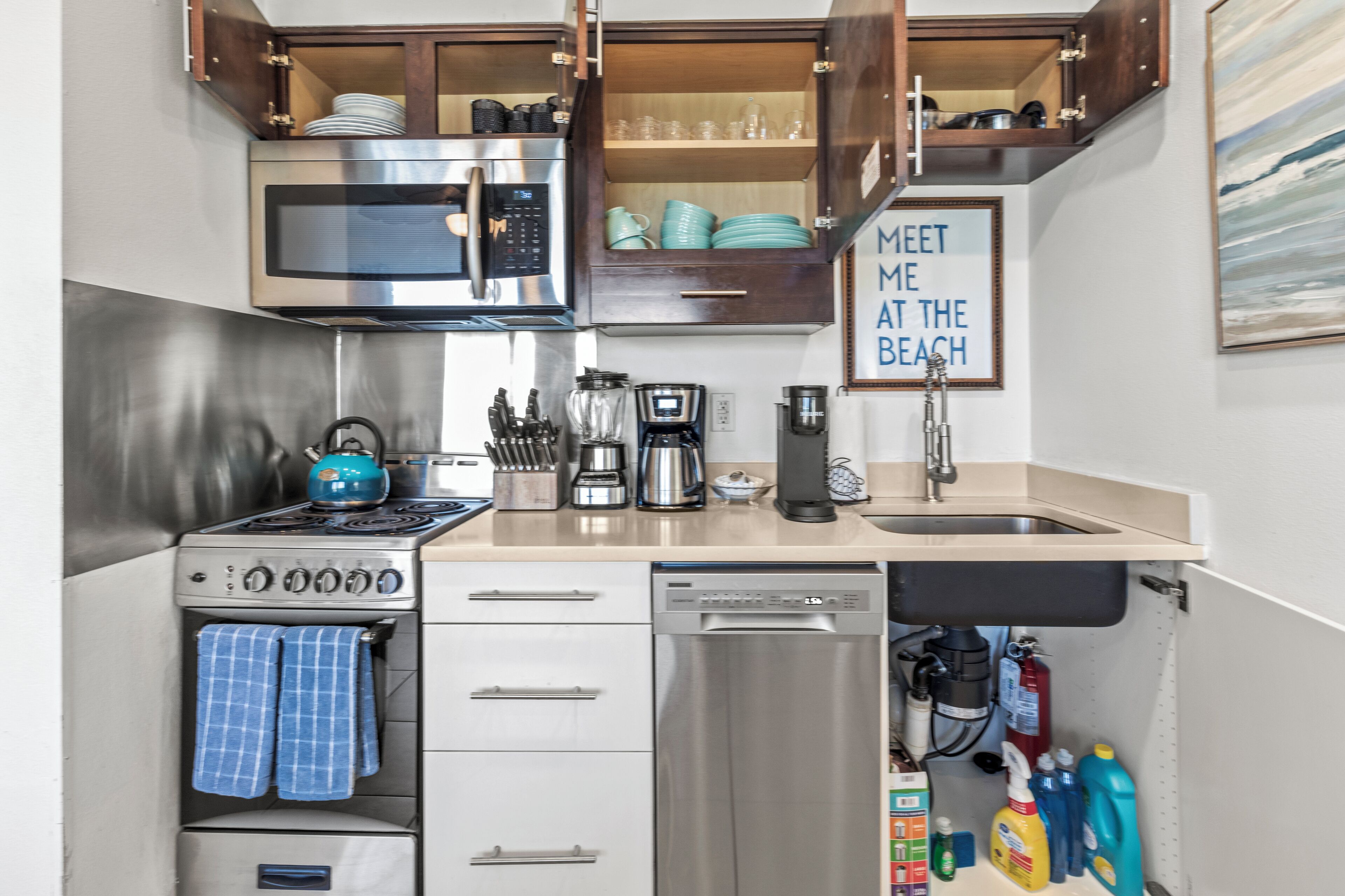 Una cocina compacta equipada con microondas, estufa y lavavajillas. Hay armarios sobre la encimera y un cartel enmarcado en la pared.