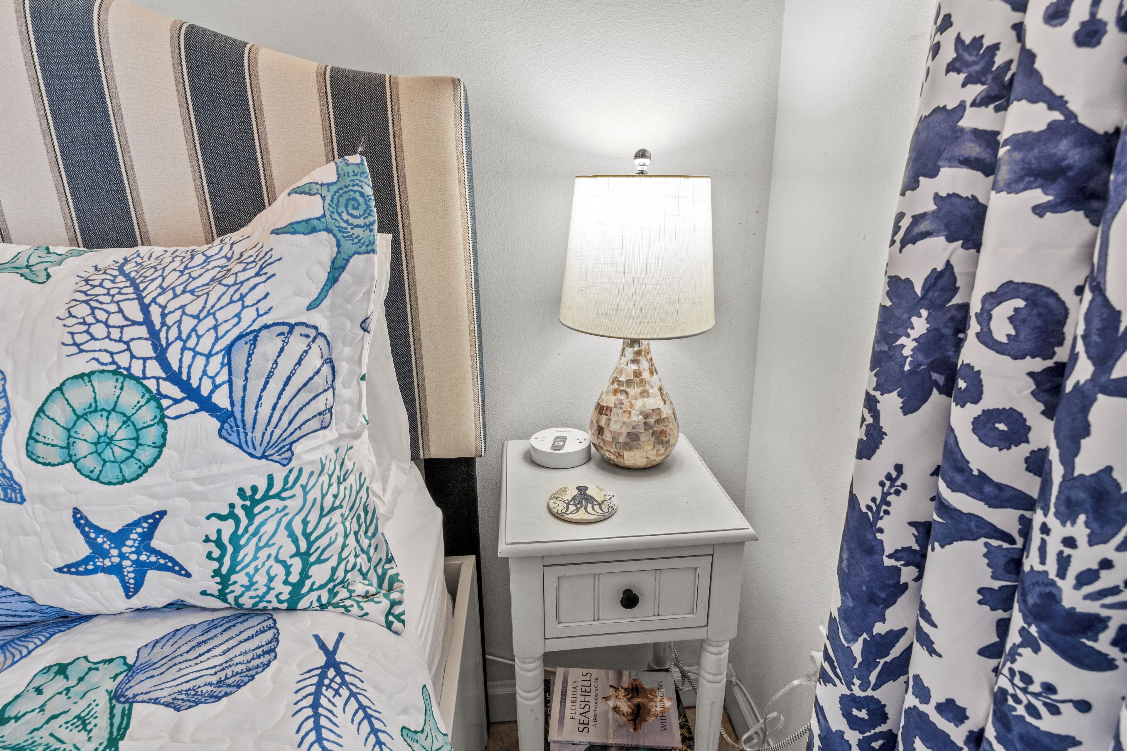 A neatly made bed with a striped pillow and a blue and white floral bedspread. A white bedside table with a lamp and a decorative item is next to the bed.