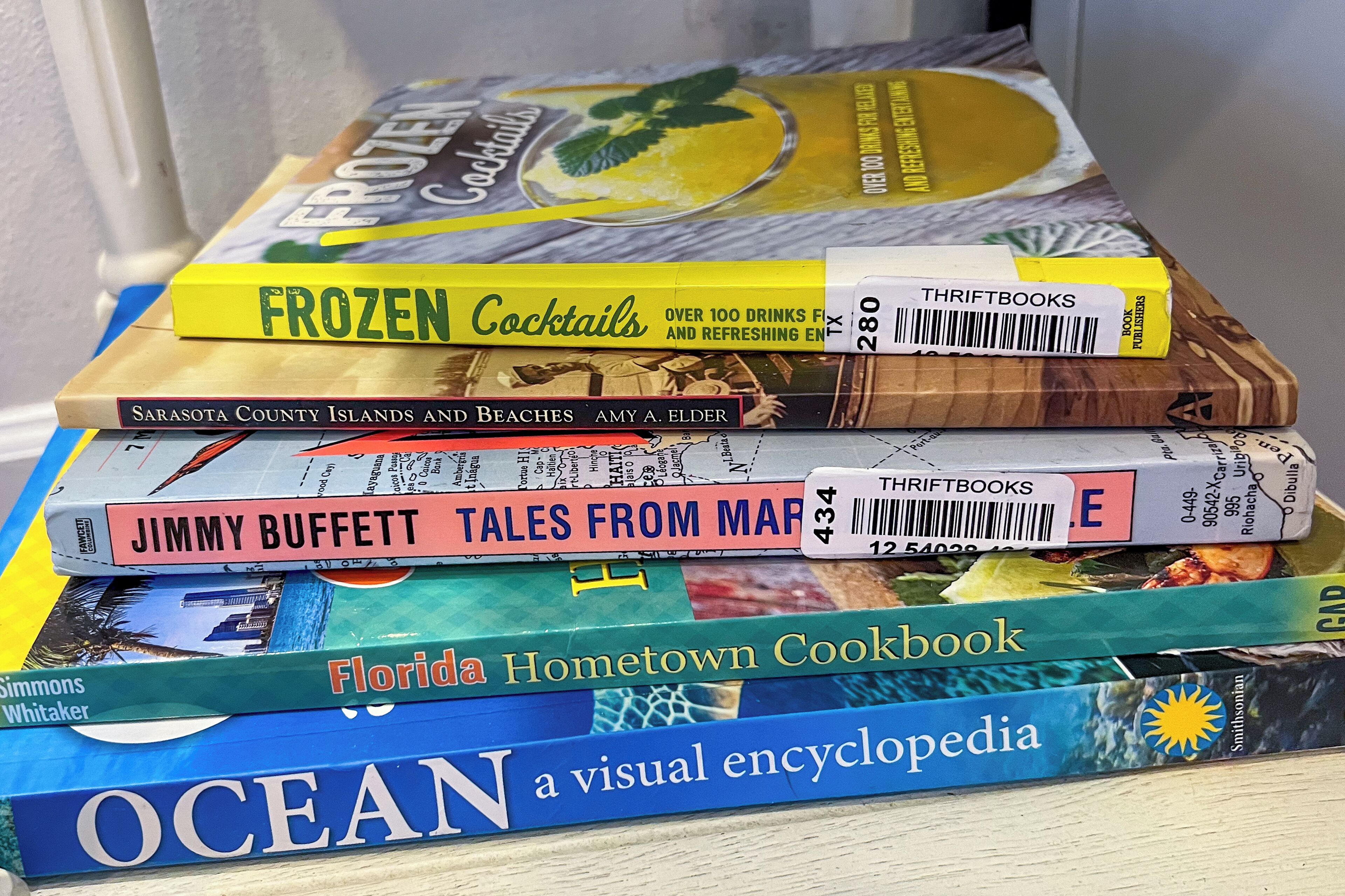 A stack of books including 'Frozen Cocktails', 'Sarasota County Islands and Beaches', 'Jimmy Buffett Tales from Mar', 'Florida Hometown Cookbook', and 'Ocean a visual encyclopedia'.