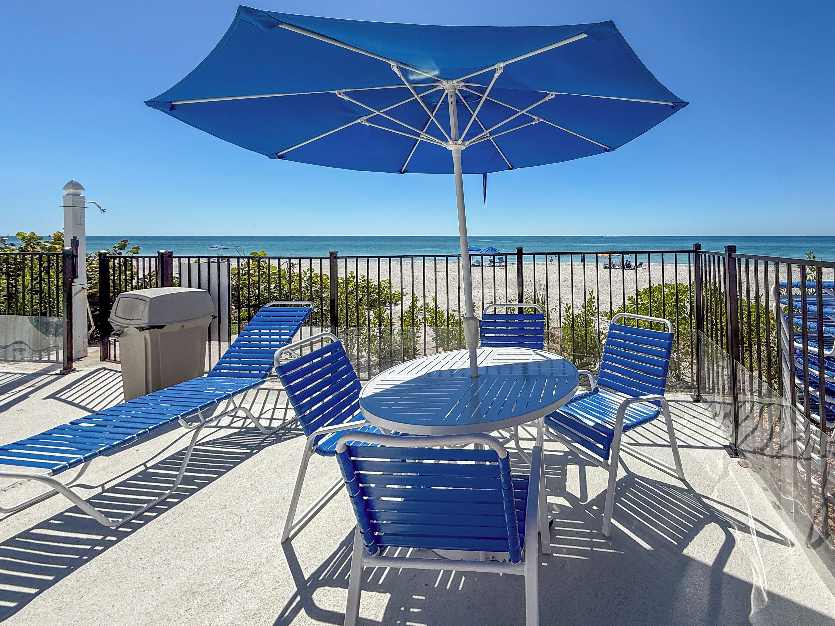 A patio with blue lounge chairs, a table, and a large umbrella, overlooking a beach.