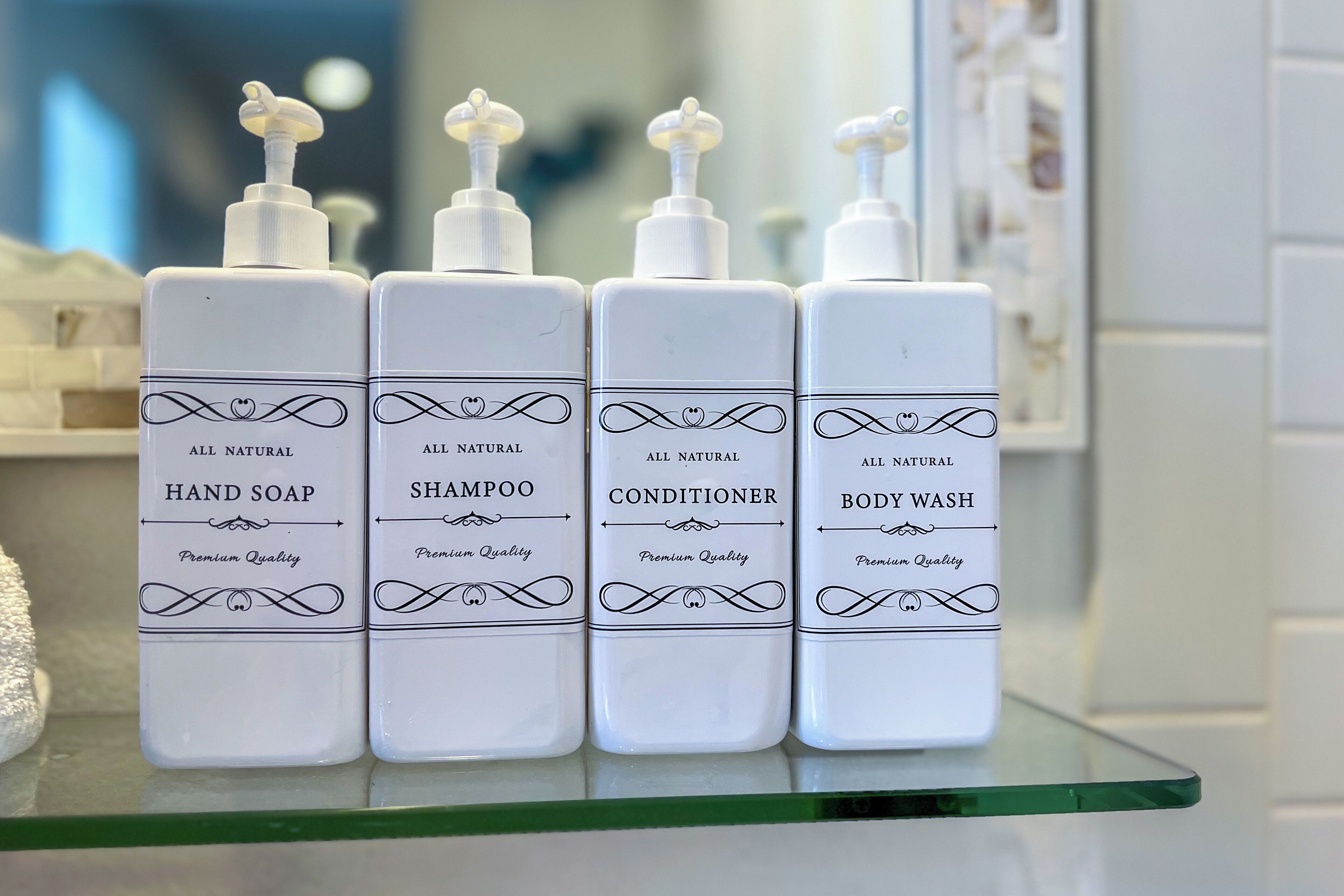 Cuatro botellas de jabón de manos, champú, acondicionador y gel de baño de calidad superior, todo natural, sobre un estante de vidrio.