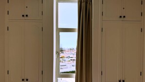 A room with a large wardrobe, a window with a view of a parking lot and buildings, and a curtain partially drawn.