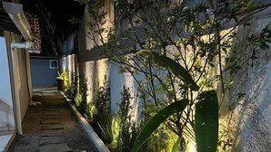 Un étroit sentier, parsemé de pierres plates, mène à l’entrée d’un bâtiment, qui est lui-même entouré d’une végétation luxuriante et de plantes en fleurs.