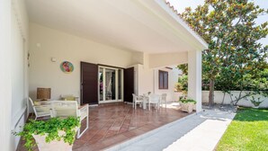 Eine überdachte Terrasse mit weißen Möbeln, ein gefliestem Boden und ein Gartenbereich mit Pflanzen und einem Baum.
