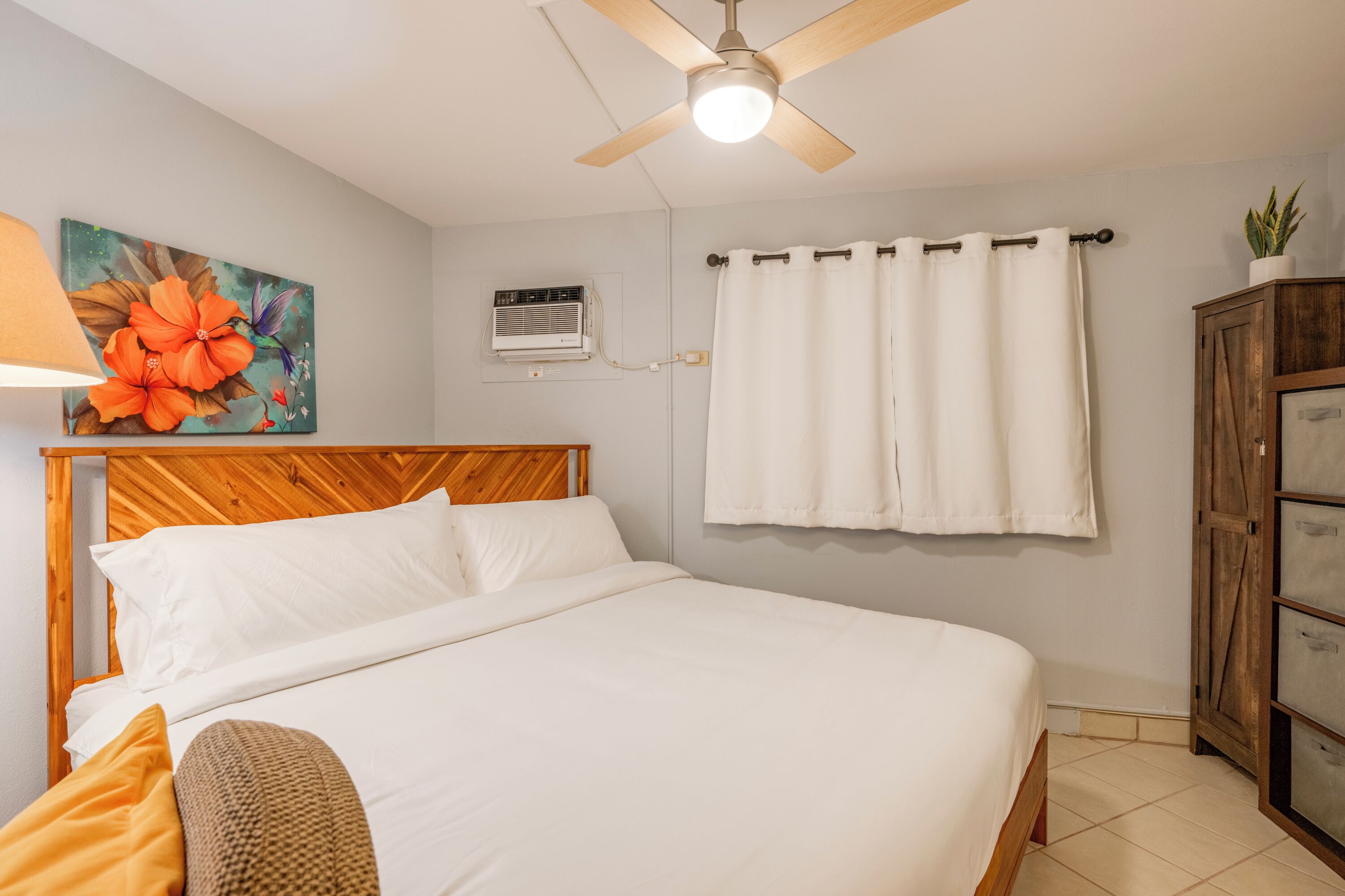 A bedroom with a wooden headboard, a bed with white linens, a ceiling fan, a wall-mounted air conditioner, a window with white curtains, and a cabinet with drawers.