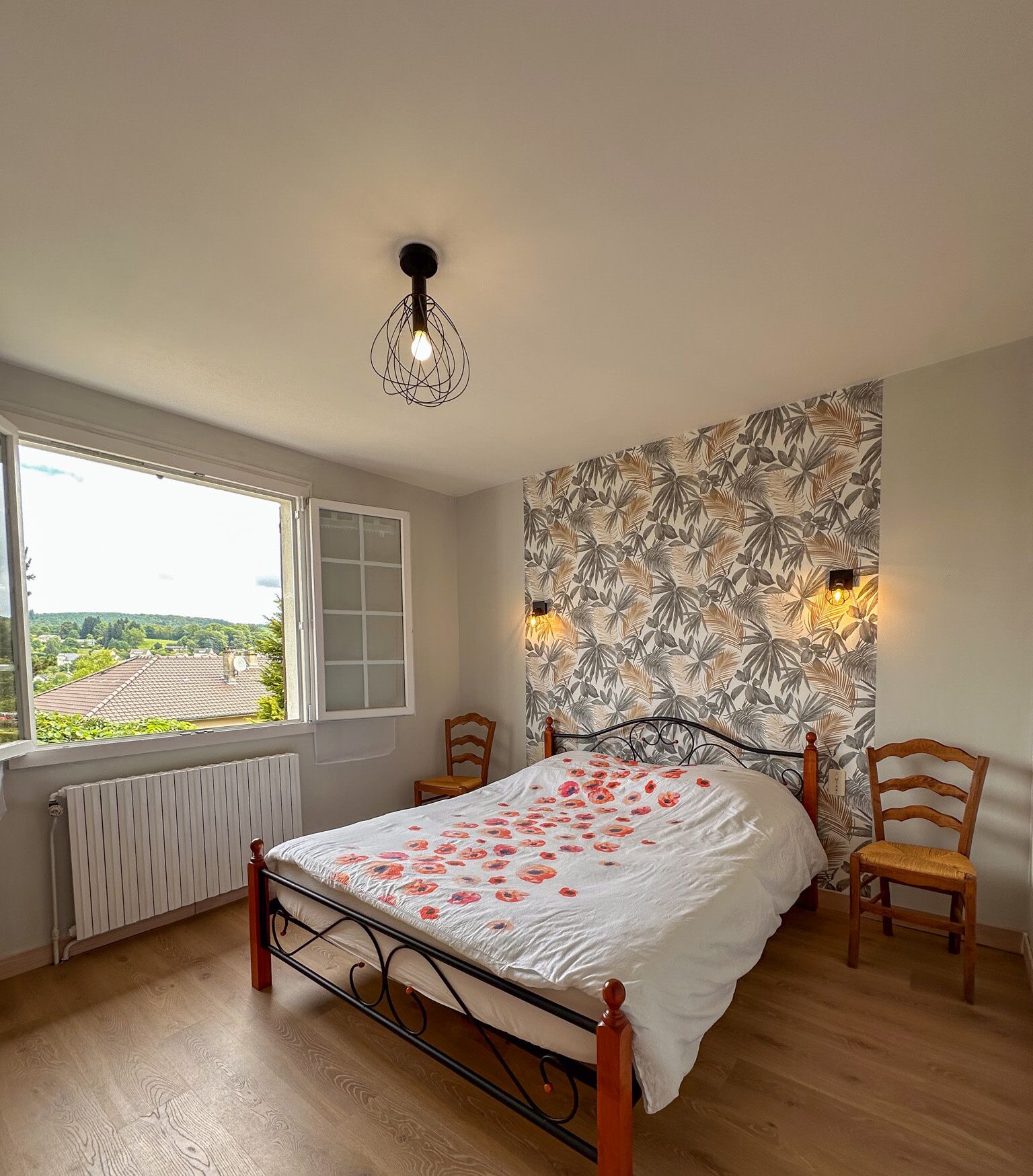 A bedroom with a bed, two chairs, a radiator, a window with a view, and a wall with a leaf pattern.