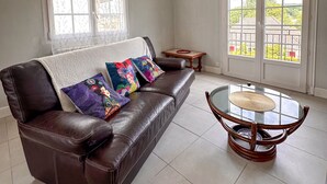 A living room with a brown leather sofa, colorful cushions, a glass coffee table, and large windows with a view of greenery outside.