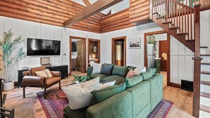 A spacious living room with a green sofa, a brown leather chair, and a flat-screen TV. A wooden staircase leads to an upper level.