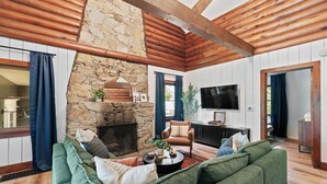 A cozy living room with a stone fireplace, green sofas, a flat-screen TV, and wooden beams.