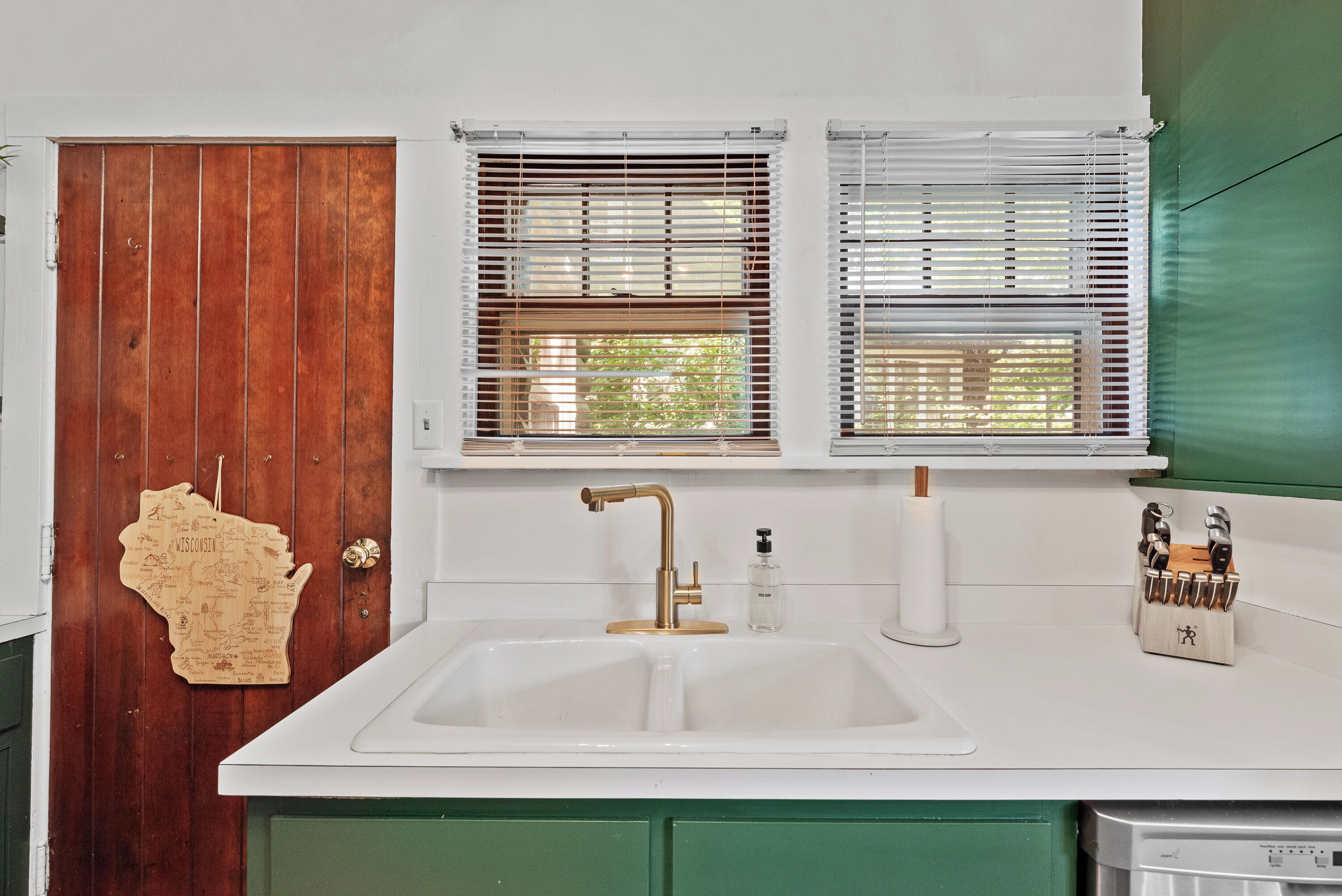 A kitchen with a white sink, two windows with blinds, a wooden door with a map decoration, and a green cabinet.