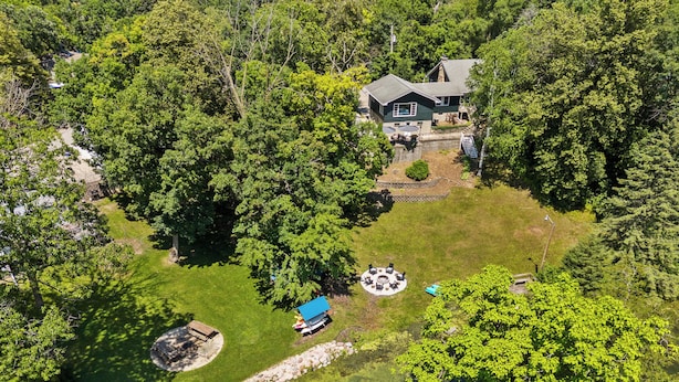 An aerial view of a house surrounded by dense trees, with a clear lawn, a circular outdoor seating area, and a small pond.