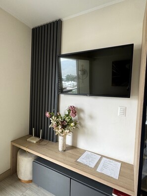 Une chambre moderne avec un téléviseur à écran plat fixé au mur, une console en bois ornée d’un vase de fleurs et un petit bureau avec un ordinateur et des documents.