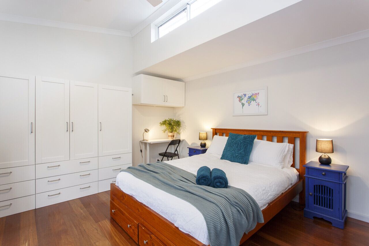Un dormitorio con cama de madera, armario blanco, escritorio y una planta pequeña.
