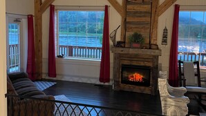 A spacious living room with a fireplace, wooden beams, and large windows overlooking a scenic view. - Island View Cottage: Waterfront-BBQ-Sauna-AC-Wifi, Island View Cottage (Flatlands)