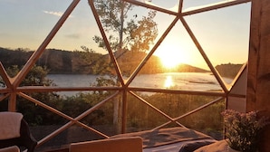 Una habitación con estructura de madera geométrica, vista a un atardecer sobre un cuerpo de agua, y una zona de estar con cojines y una planta.